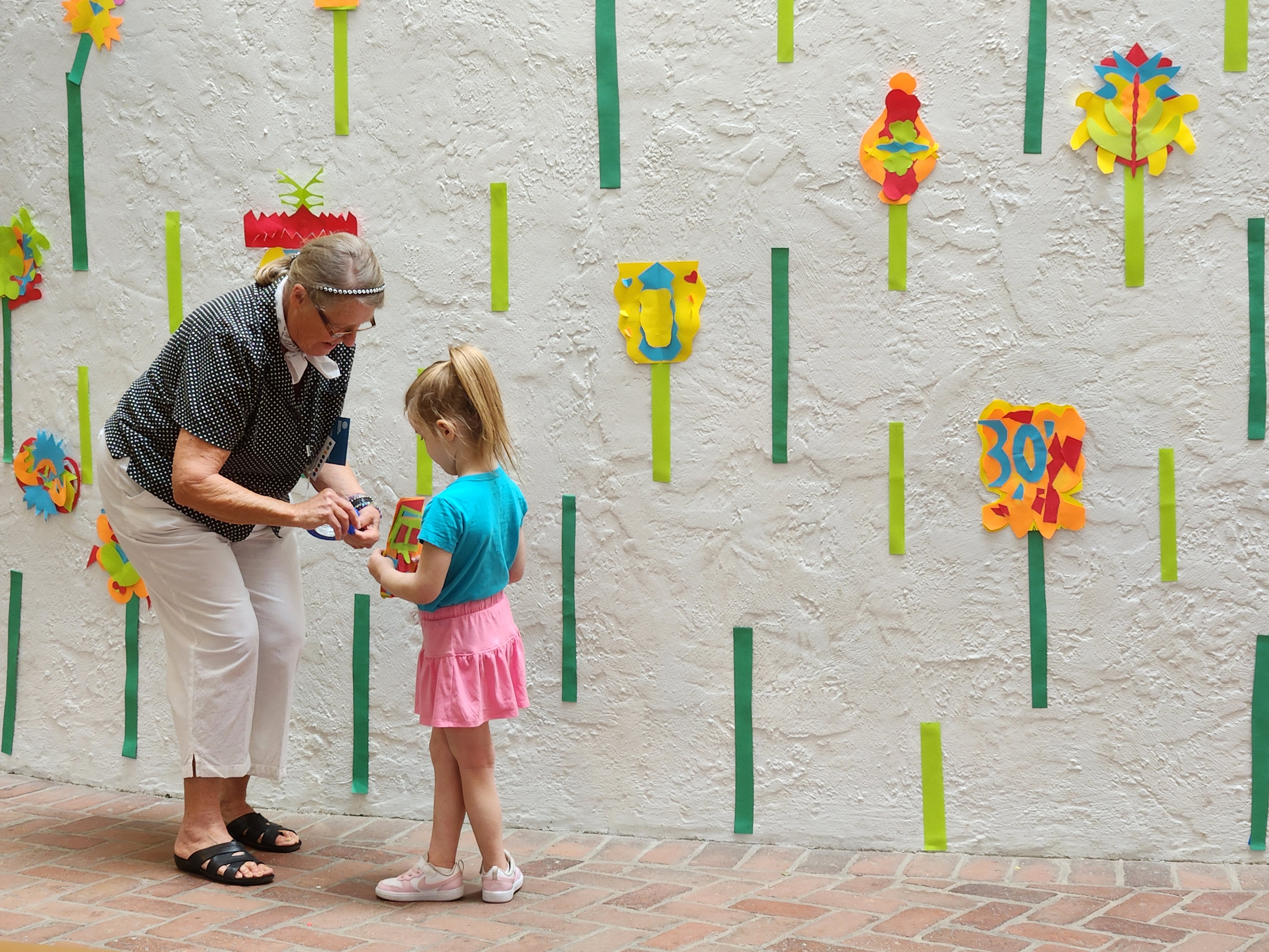 A woman and young girl stand against a colorful, crafted wall talking.