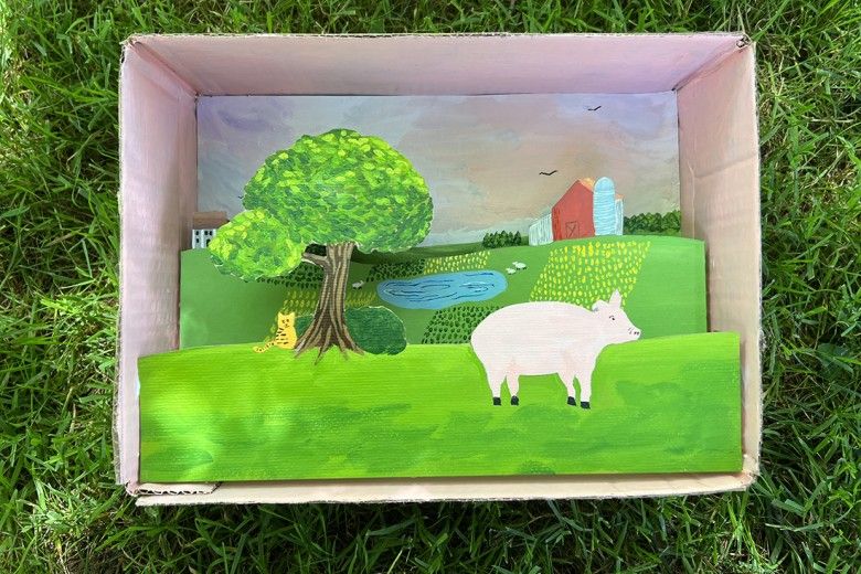 A finished landscape shadowbox example. This photo includes an open cardboard box that is filled with a colorful scene featuring a pig on a farm landscape.