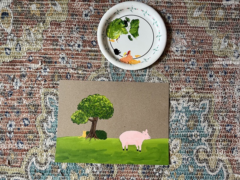 A pink pig on a green, grassy field with a tree in the background painted on a thin piece of cardboard. Above the painting is a white paper plate with globs of paint on it.