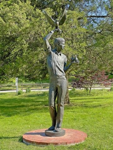 Outdoor bronze sculpture of a larger-than-life young man holding two doves in one hand and a hawk in the other.