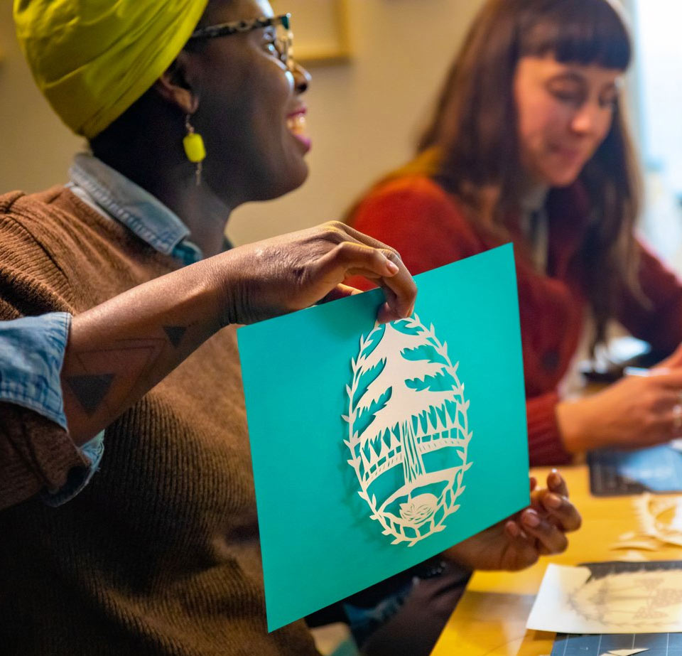 A woman holding up a paper cut artwork with another woman in the background.