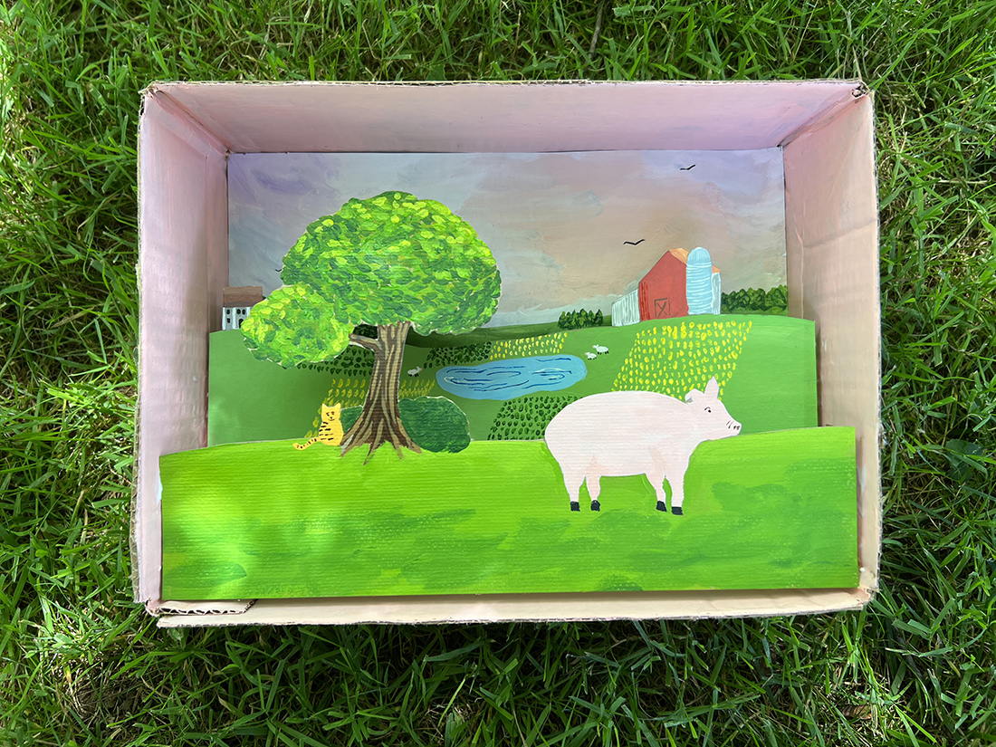 A finished landscape shadowbox example. This photo includes an open cardboard box that is filled with a colorful scene featuring a pig on a farm landscape.
