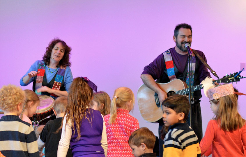 Two musicians playing guitar and drums surrounded by kids.