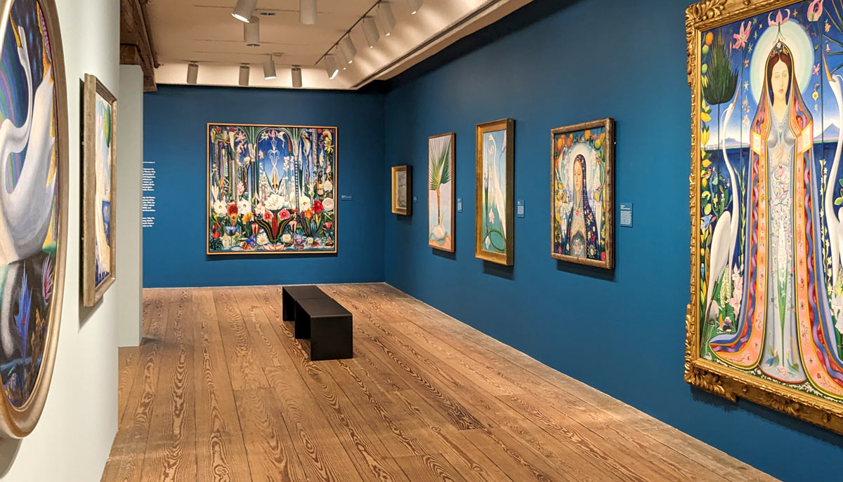 An empty art gallery with paintings hanging on blue walls and a gray bench in the middle of the room.