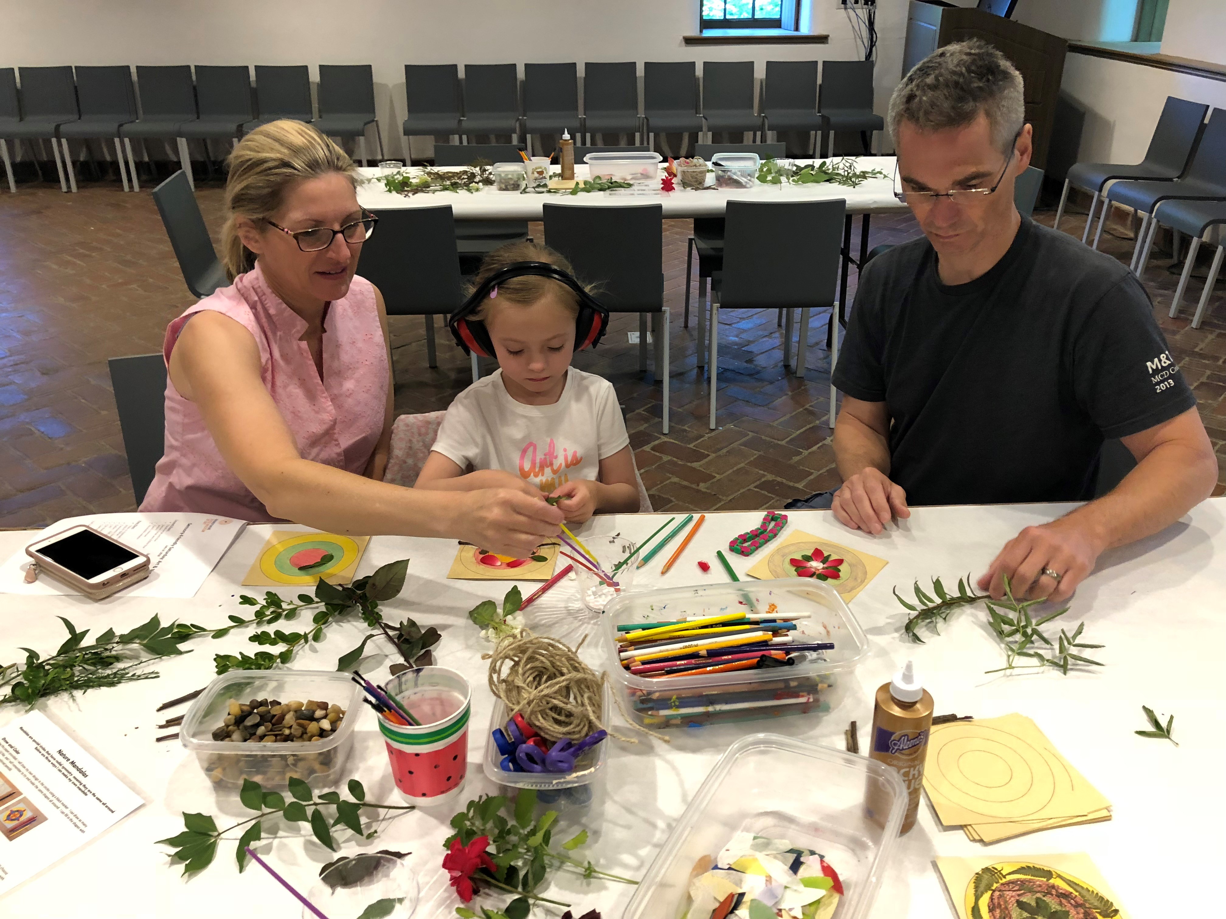 A child sits with her guardians at a table doing crafts with objects of nature.