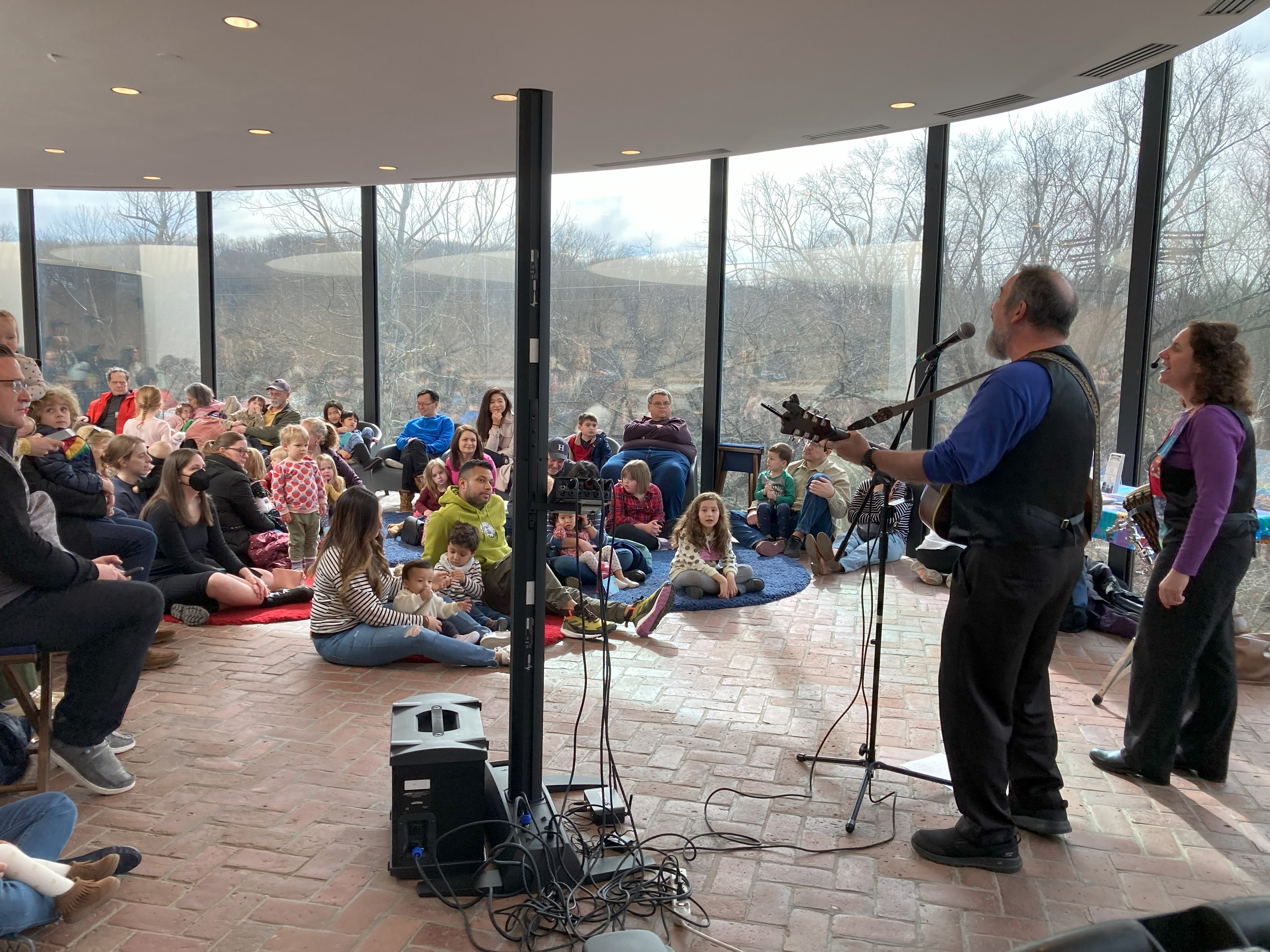 A man plays the guitar and sings in front of children and their families who are sitting on the floor watching.