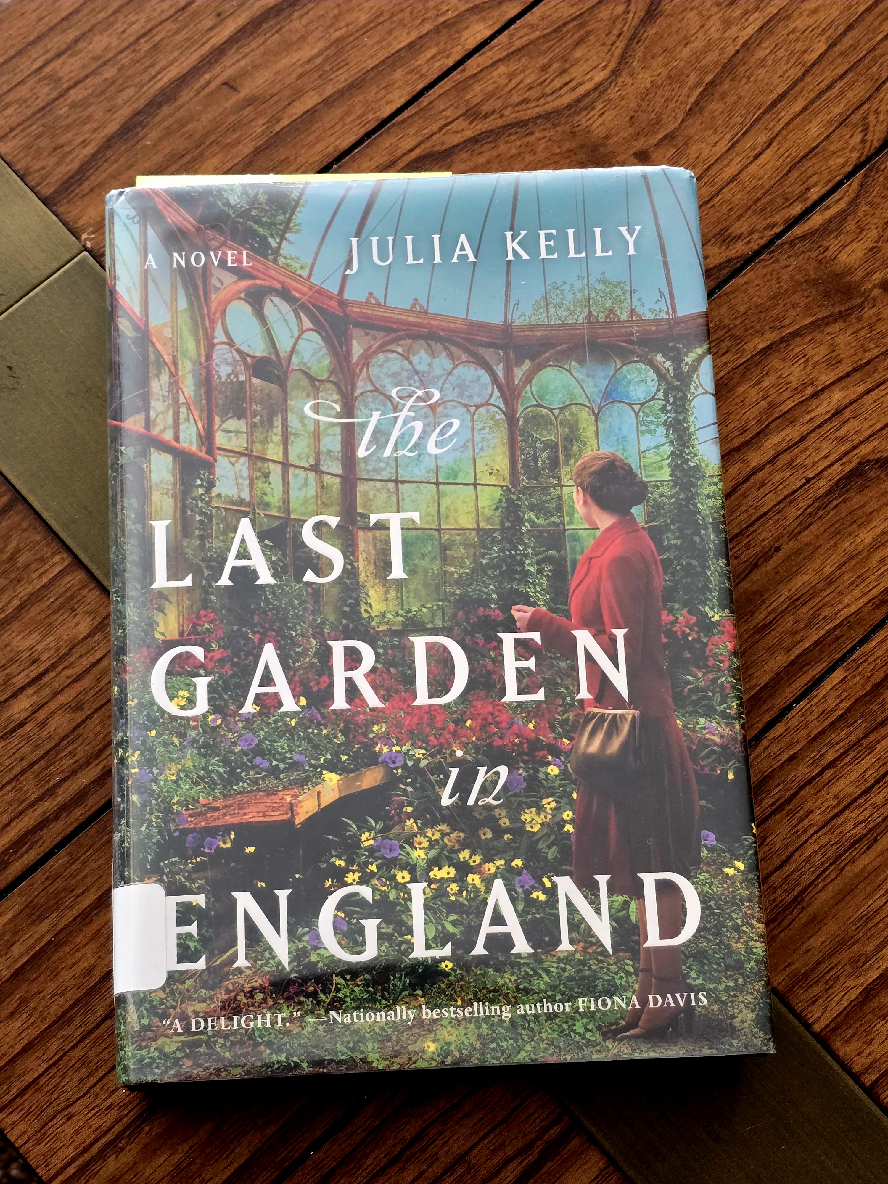 A book resting on a table with a women in a green house on the cover with white words reading The Last Garden.