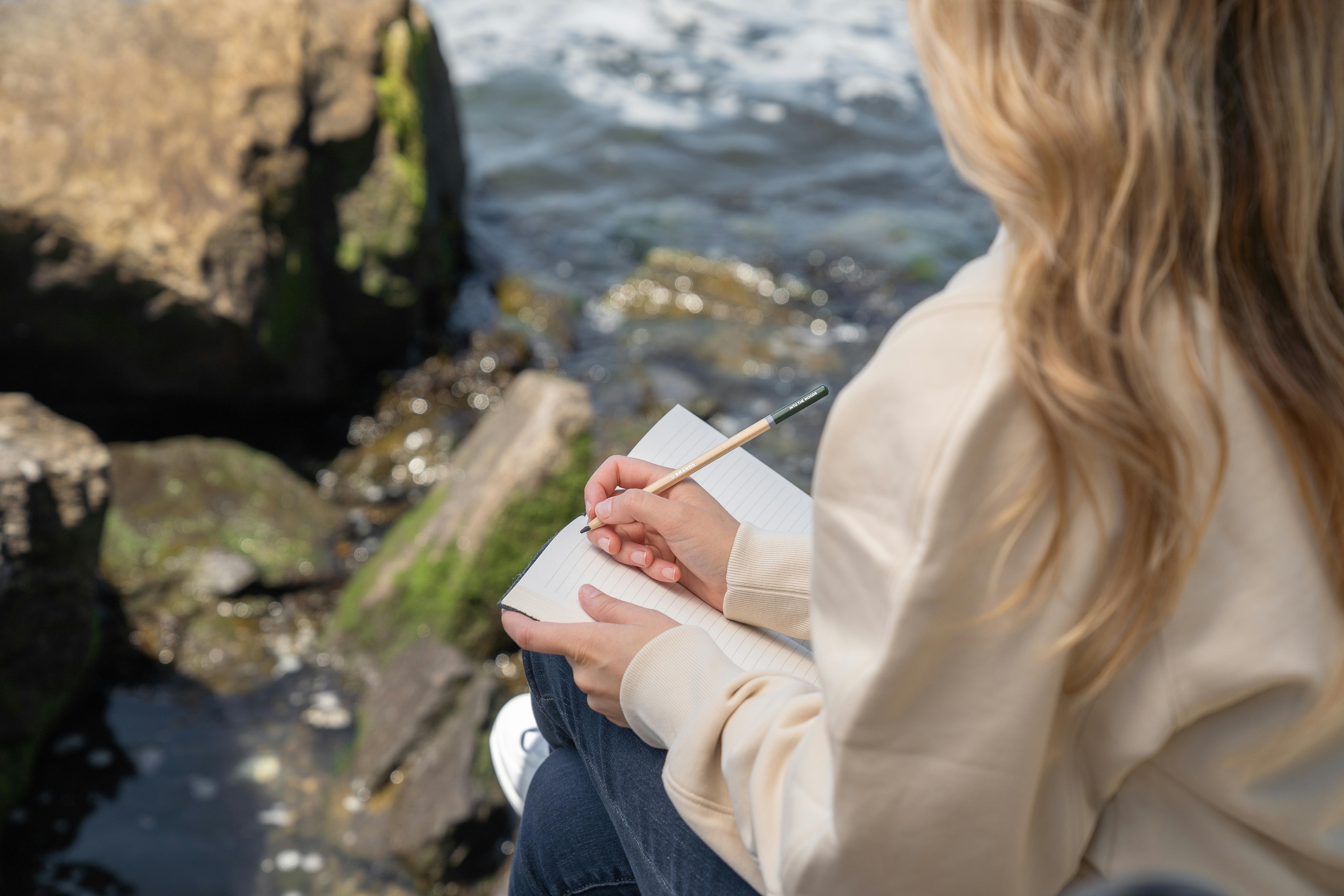 A girl sits by the water with an open journal.