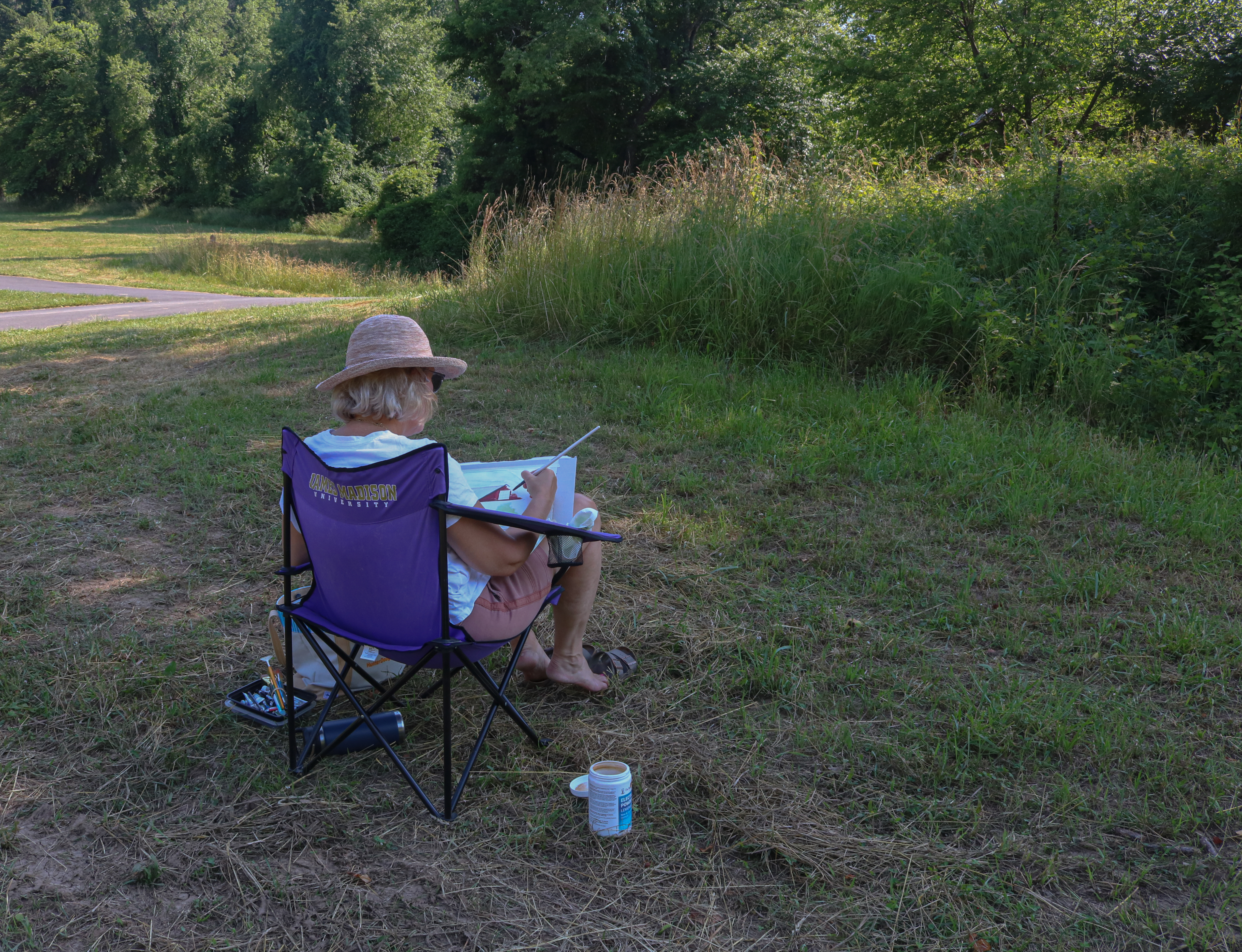 A women sits in a lawn chair with paint materials in an open, grassy field.