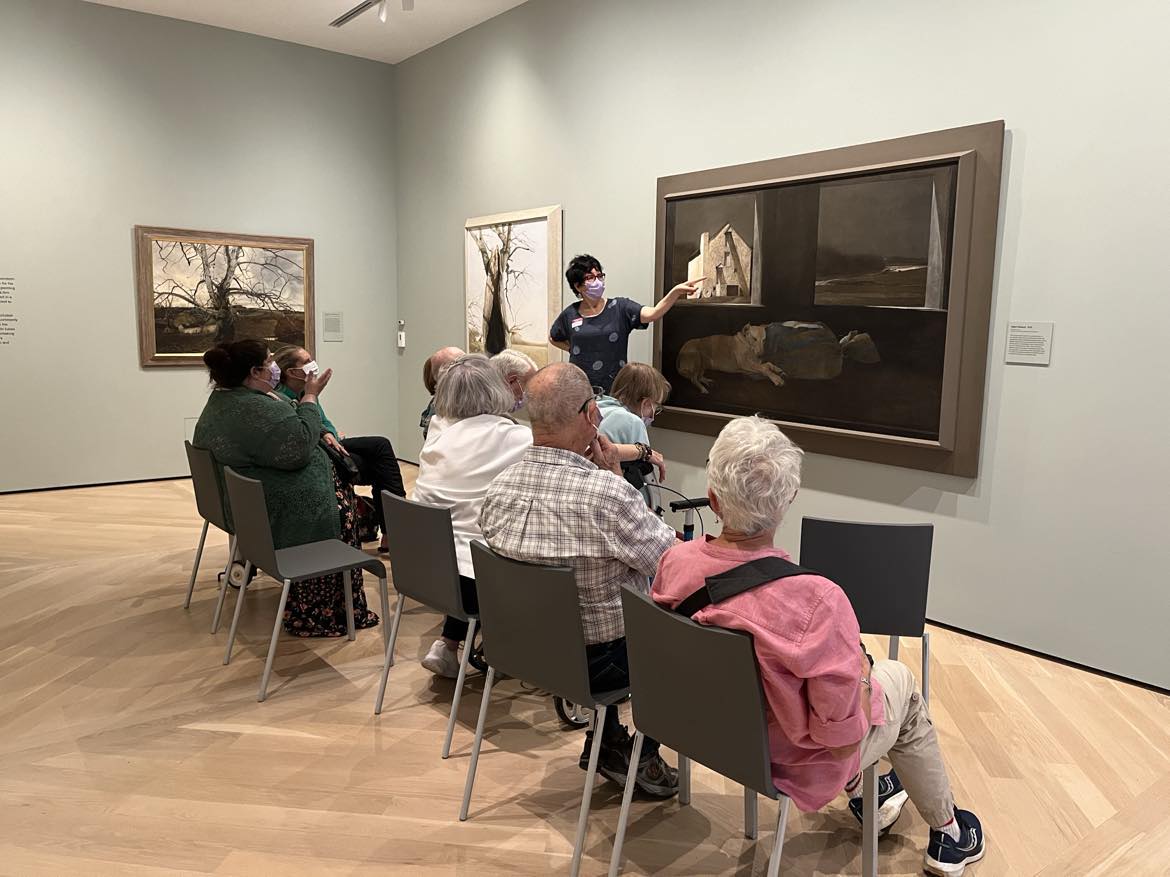 People sitting in chairs looking at a painting, while someone talks about the painting