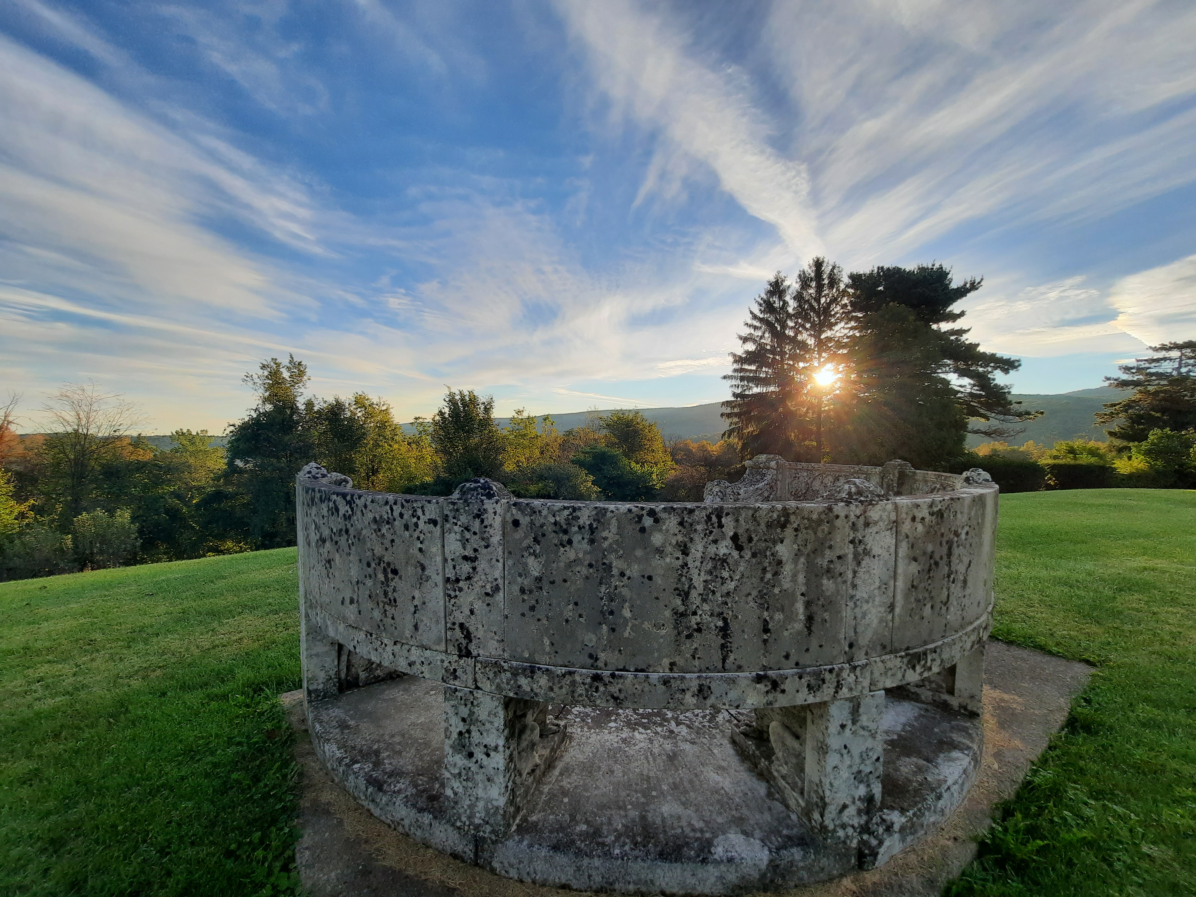 A curved stone bench situated on top of a hill, surrounded by trees and the setting sun.