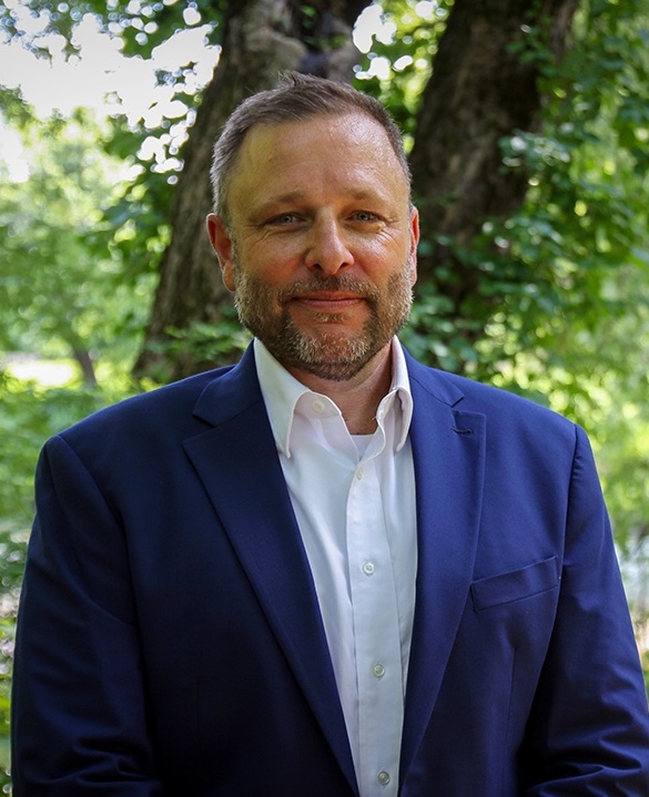 Headshot of Grant DeCosta, pictured outside in a blue suit and white shirt, with a tree behind him.
