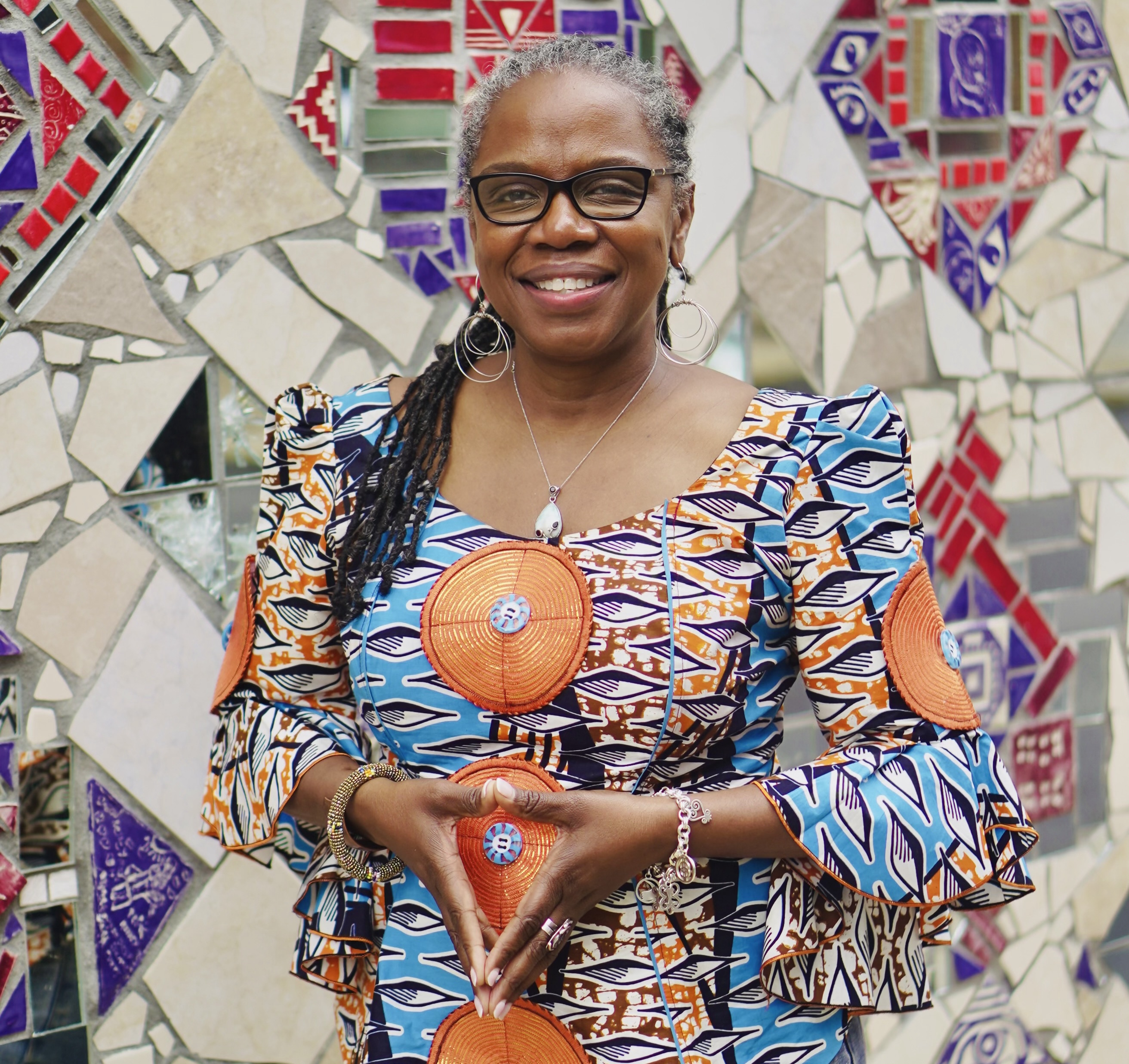A woman in a bright patterned dress, standing in front of a mosaic wall