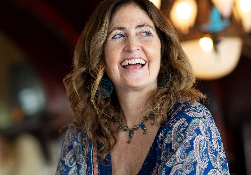 Photo of A woman with light skin and wavy shoulder-length brown hair is smiling and looking off to the side. She’s wearing a blue patterned top, earrings, and a necklace. The background is softly blurred with warm lights.
