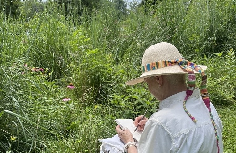 A woman wearing a straw hat with a multicolored ribbon on it. She is facing away from the viewer and drawing on a pice of paper while looking at a field of tall grass with purple flowers sprinkled throughout.