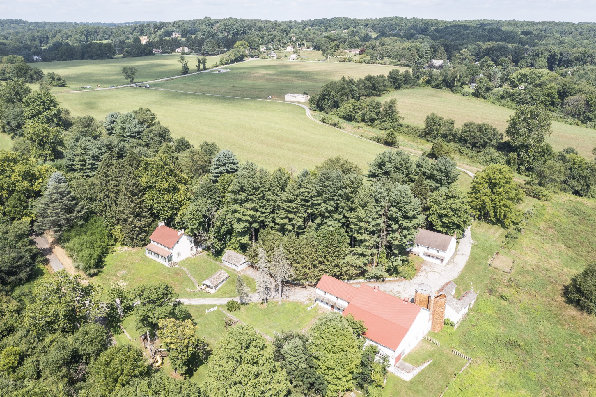An aerial view of a quiet rural property surrounded by wide, open fields and dense green trees. In the foreground, several buildings form a small farmstead: a white farmhouse with a red roof, a large barn with a red metal roof and two tall silos, and a few smaller outbuildings connected by a curved driveway.