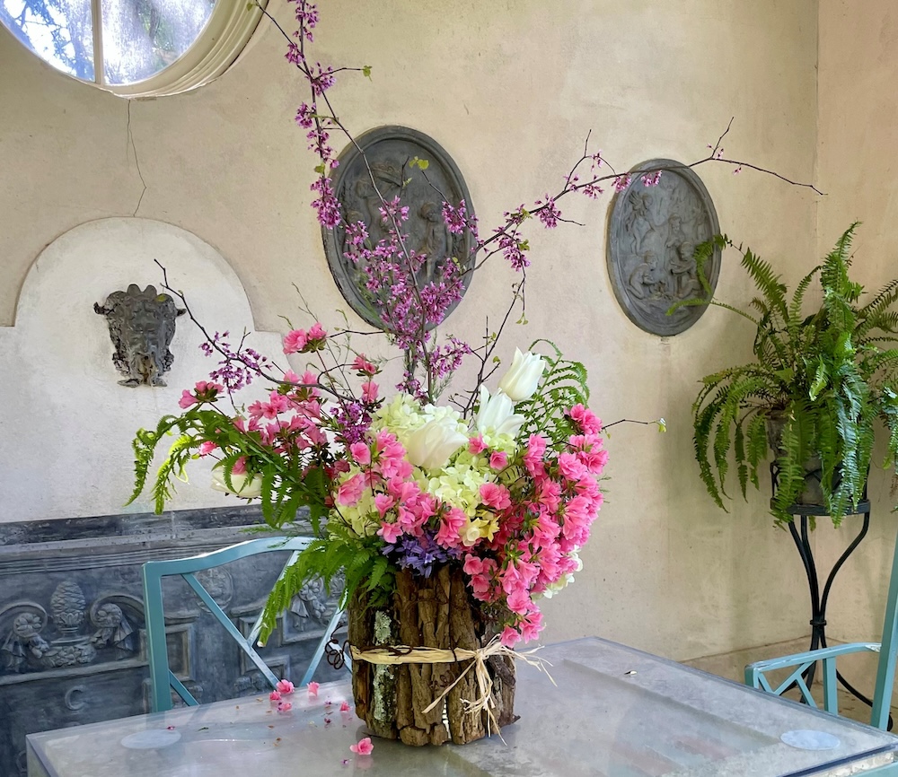 A rustic wooden vase tied with twine sits on a metal table, overflowing with pink blossoms, soft white flowers, and airy branches that reach upward and outward. Behind it, a pale stucco wall with carved stone plaques, a small round window, and leafy ferns creates a quiet garden-like setting that feels calm.