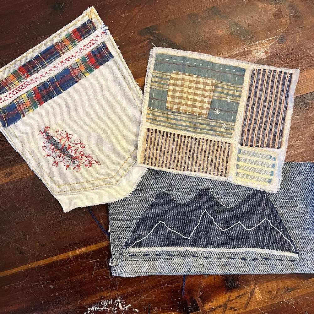 Photo of piece of fabric laying on a brown wooden table. Each piece of fabric has been hand stitched with threads of various colors.