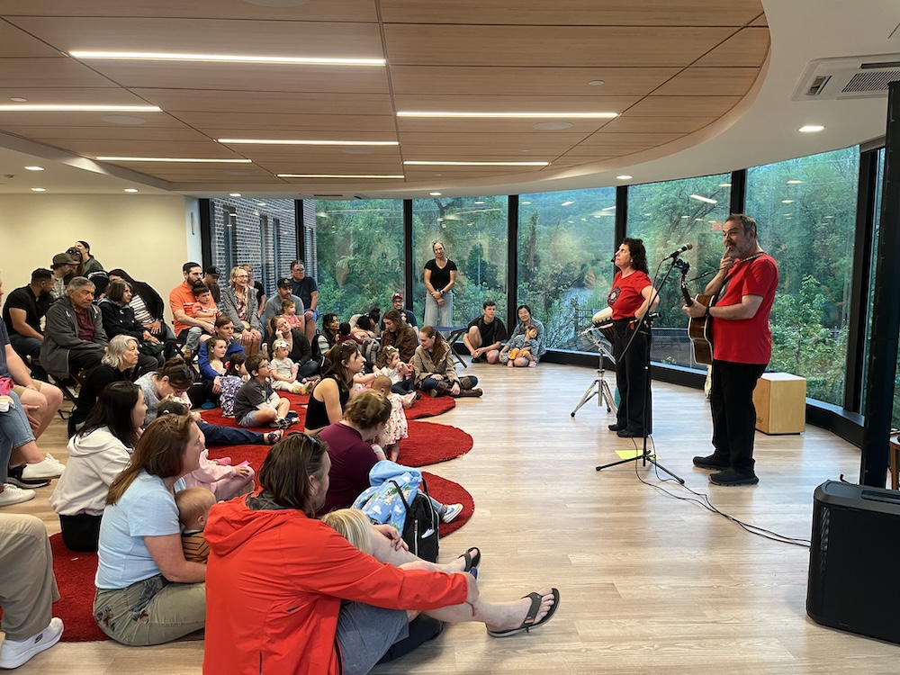 A group of families with many young children sit closely together on the floor and chairs, watching two performers at the front of a bright room with large windows overlooking greenery. One performer sings into a microphone while the other plays guitar, creating a small, intimate live music experience for the audience.
