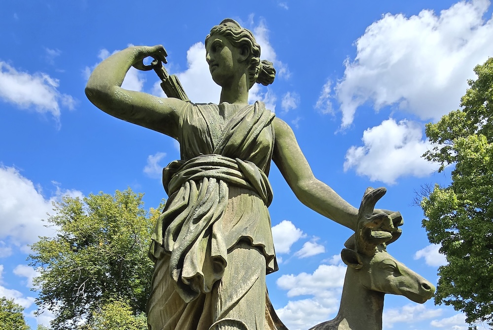 A classical-style statue of a woman stands outdoors against a bright blue sky, holding a bow over her shoulder and resting one hand on a deer beside her. The sculpture is surrounded by green trees, giving it a serene, natural setting.
