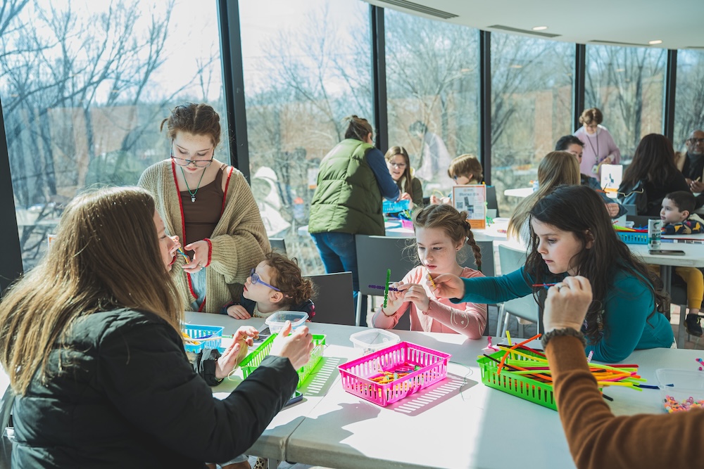 A group of children and a few adults are gathered around tables in a bright room with large windows, working on colorful craft activities together. They are using items like pipe cleaners and small tools, focusing closely and interacting as they create their projects.