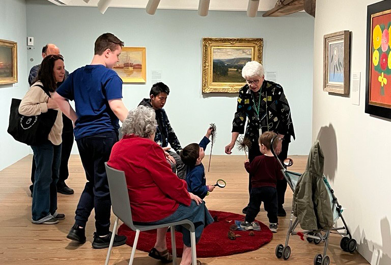 A museum educator leads a tour with children and adults in a gallery, on a red mat surrounded by landscape and still life paintings.