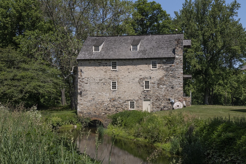 An old stone mill sits beside a peaceful stream, surrounded by lush green trees.