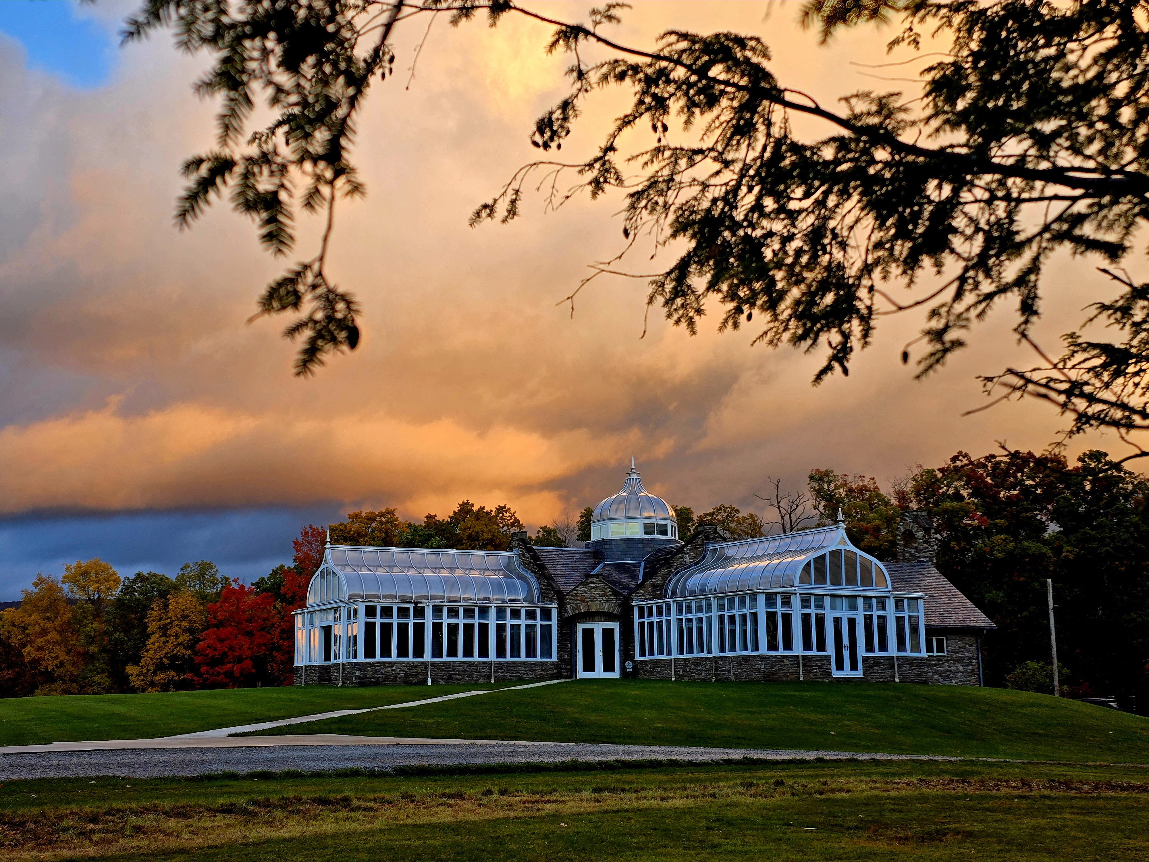 Clouds backlit by the sun, a glass greenhouse in the foreground with framing the building