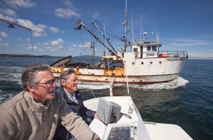 Two men on a boat looking put at the water, with a larger boat in the background