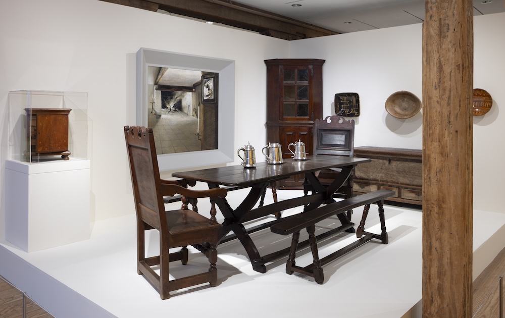 A museum exhibit displays antique wooden dining furniture and household objects.