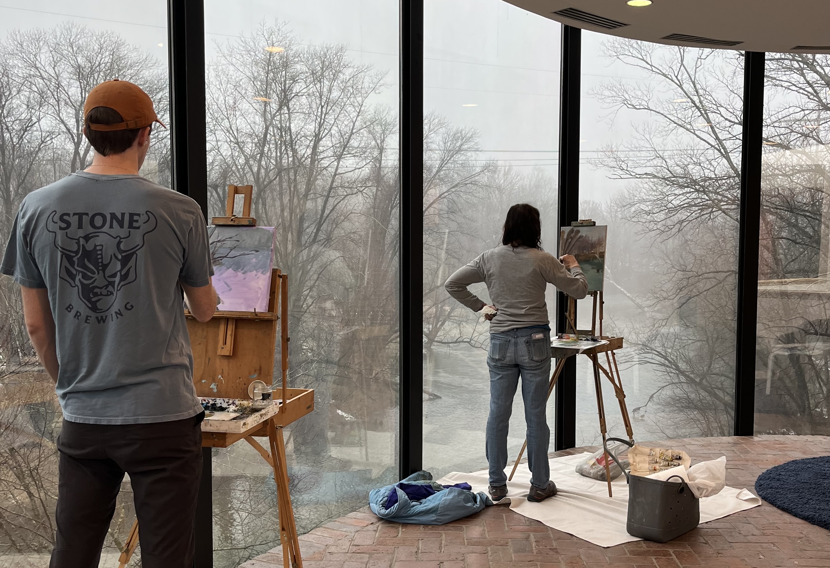 Two people paint at easels in a modern, glass-walled room overlooking a misty landscape, with art supplies spread across protective drop cloths on the brick floor.