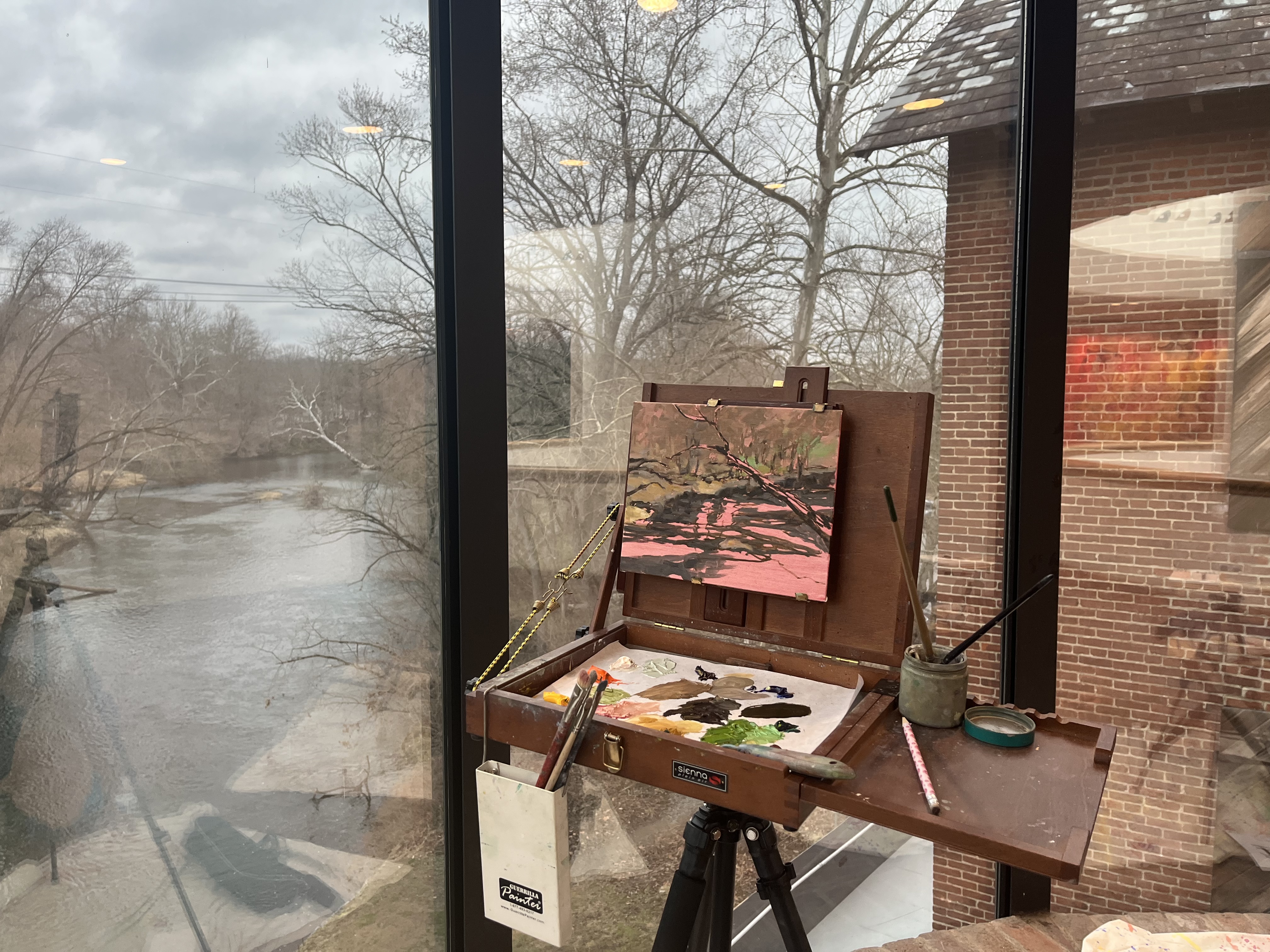 An artist’s easel and painting setup sits beside a large window overlooking a quiet river and leafless trees on a cloudy day.