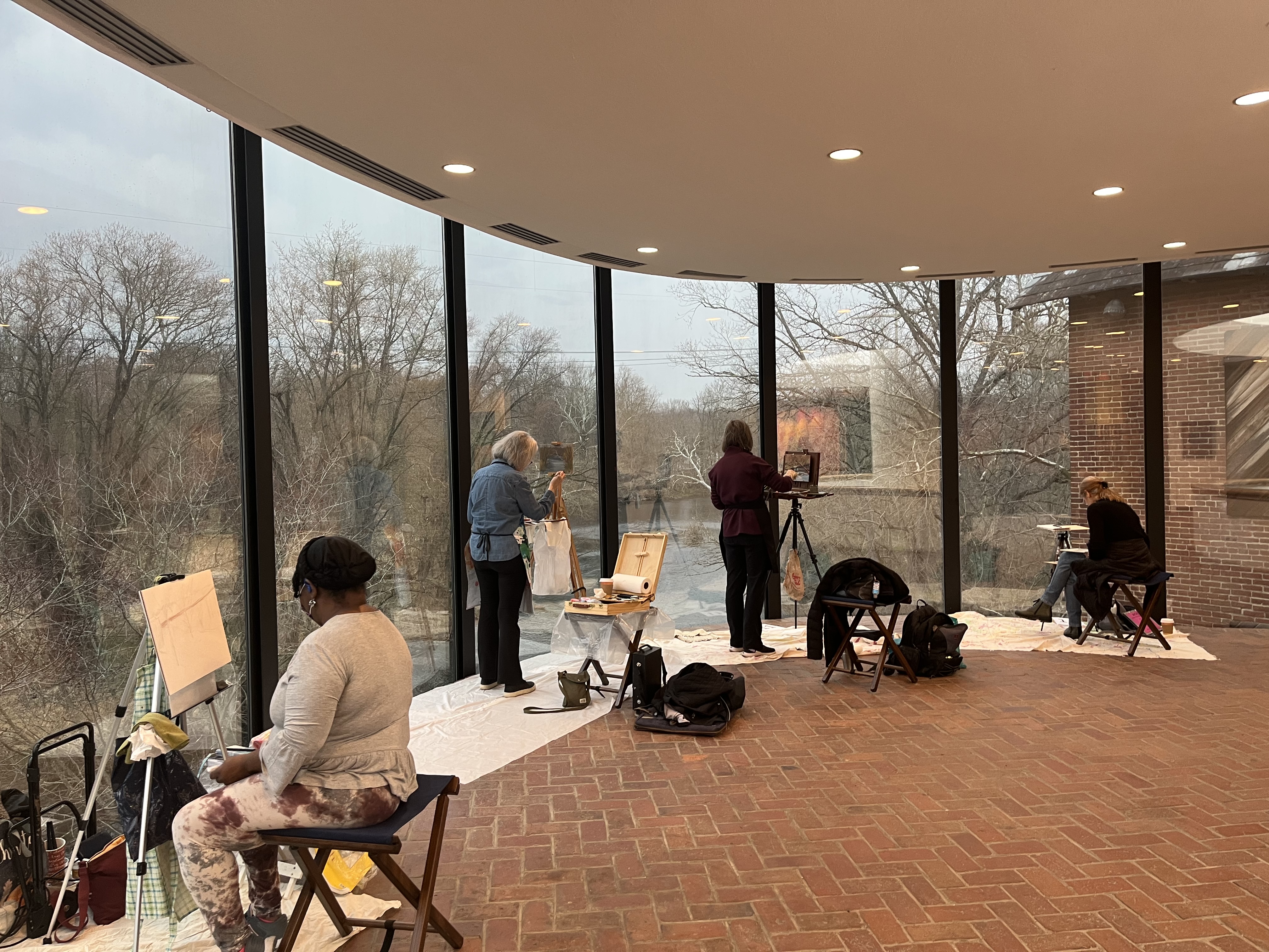 Several artists sit by floor-to-ceiling windows in a brick-floored room, painting and sketching the leafless trees and peaceful landscape outside.