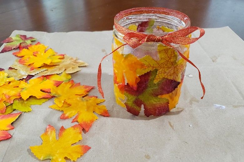 Glass candle holder covered in fall leaves, with an orange ribbon wrapped around the top, sitting on a table with more fall leaves scattered around it
