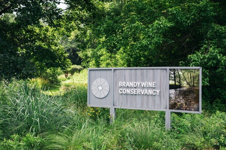Brandywine Conservancy logo on a grey wooden sign situated outside in a lush, green landscape