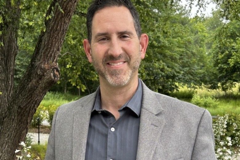 a man in a grey suit standing in front of lush green trees and flowers