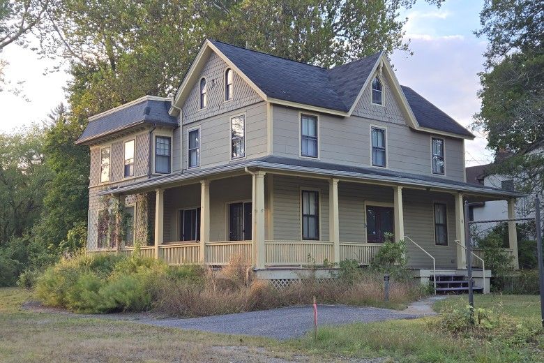 A two-story house, with a dark roof and a full wraparound porch painted in a light tan color. The exterior siding is a muted greenish-gray. The house is surrounded by overgrown trees and brush in the late afternoon light.