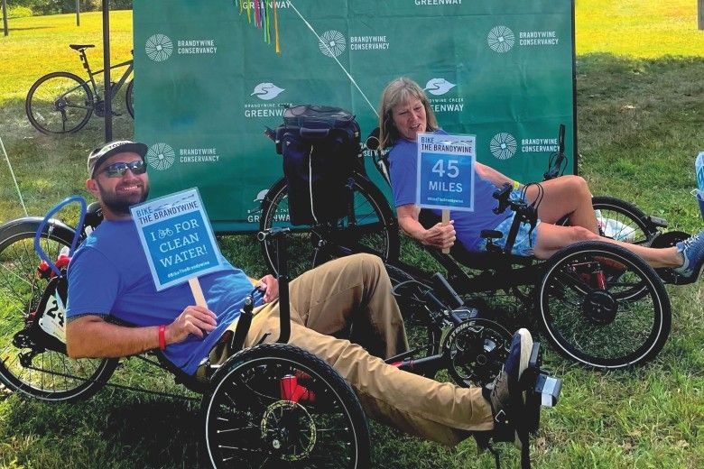 Two people are relaxing on low, three-wheeled recumbent bicycles at an outdoor community event on a sunny grassy field. They are smiling and holding signs promoting a cycling fundraiser for clean water and the Brandywine Greenway, with one sign showing a 45-mile ride option.