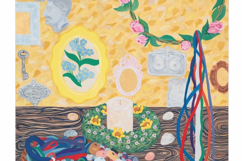 A colorful still life shows a table filled with ribbons, beads, shells, and fruit, with a candle in a flower wreath at the center. Behind it, a bright yellow wall is decorated with frames, sculpted faces, flowers, and hanging ribbons.