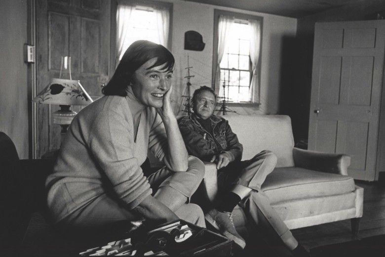 A black-and-white photograph shows two people relaxing in a cozy living room, with one person seated in the foreground smiling and resting their head on their hand. The other person sits casually on a couch behind them, surrounded by a lamp, windows, a model ship, and simple wooden furnishings.