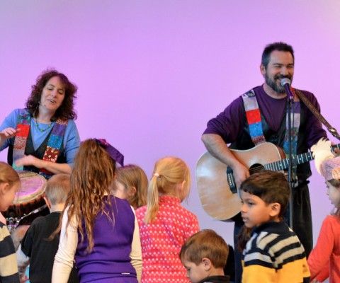 Two musicians playing guitar and drums surrounded by kids.