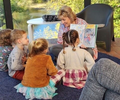An older women sits on a chair reading a book to a group of younger children who are sitting on the carpeted floor surrounding her.