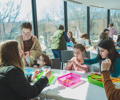 A group of children and a few adults are gathered around tables in a bright room with large windows, working on colorful craft activities together. They are using items like pipe cleaners and small tools, focusing closely and interacting as they create their projects.