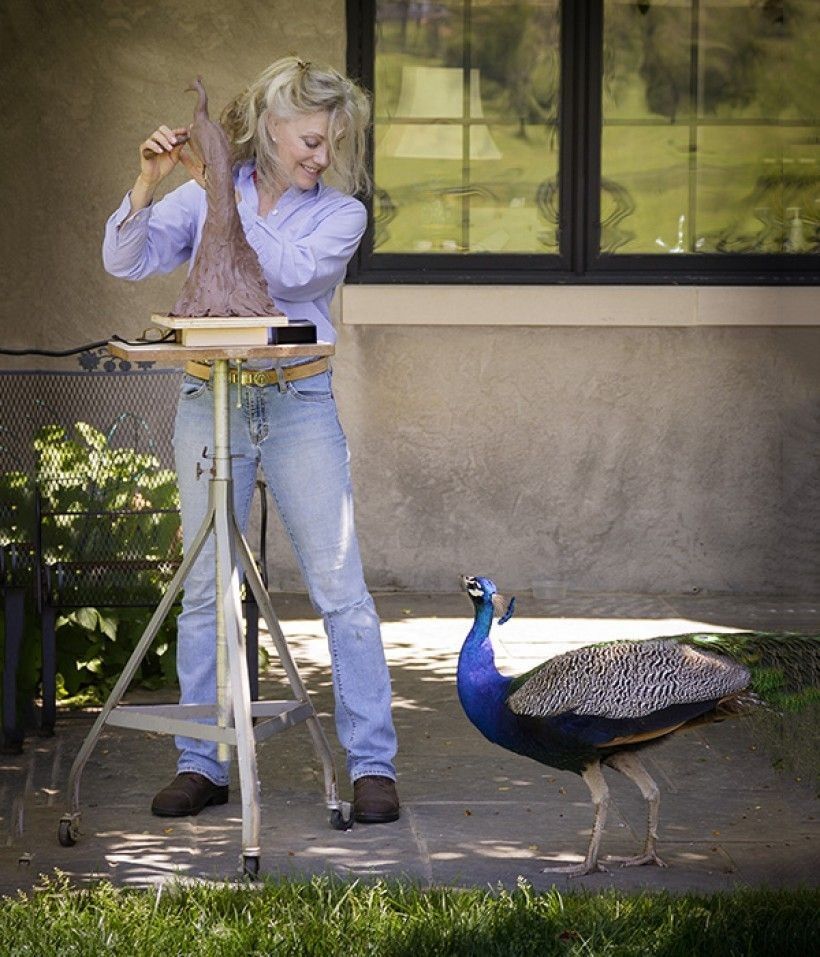 Rikki Morley Saunders photographer working on a sculpture of a peacock, with a real peacock bird at her feet