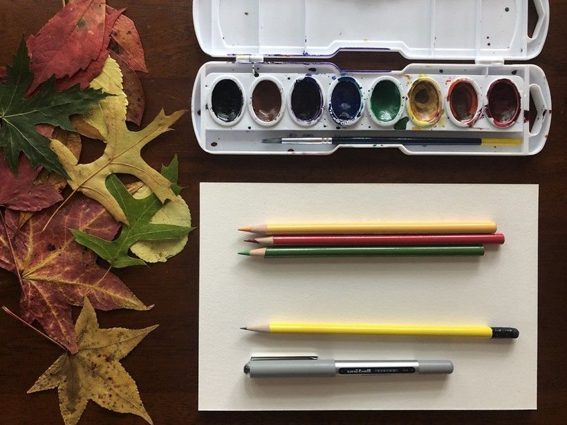 Photo of supplies needed for fall foliage designs, including: fall leaves, blank paper, watercolor paint, colored pencils, and a pen