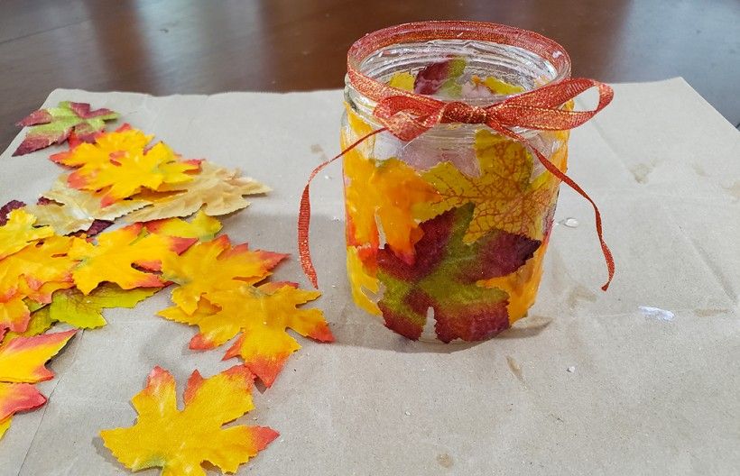 Glass candle holder covered in fall leaves, with an orange ribbon wrapped around the top, sitting on a table with more fall leaves scattered around it