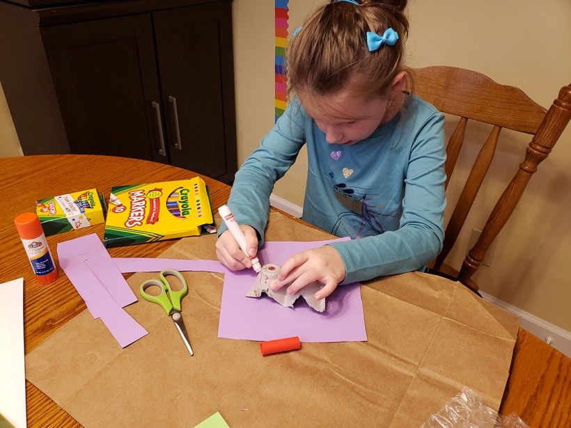 A young girl using a marker to draw eyes on a cut-up egg carton 