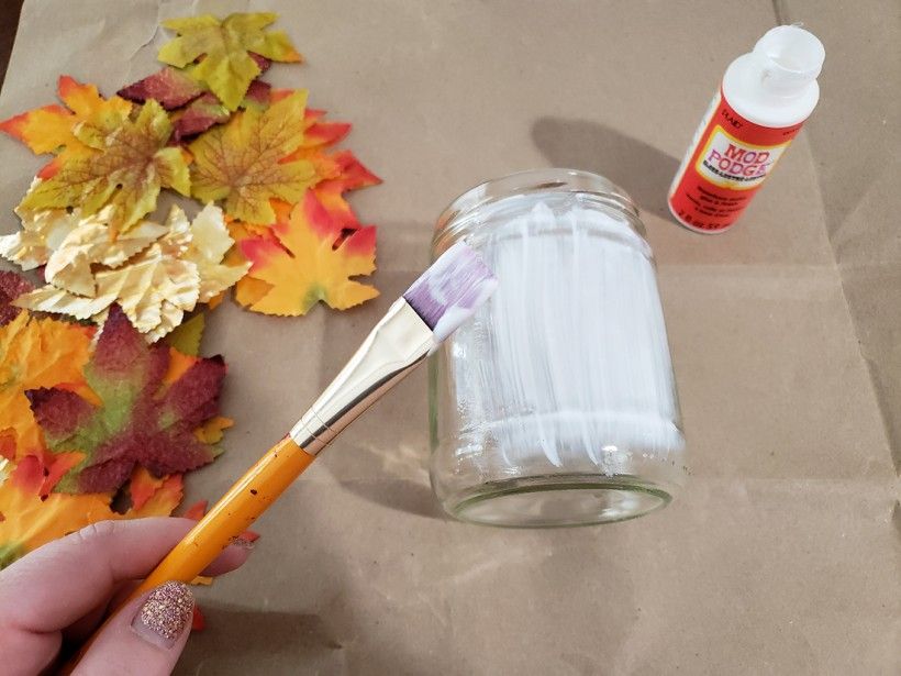 A paintbrush apply mod podge to a glass candle holder, with fall leaves and a mod podge container on a table