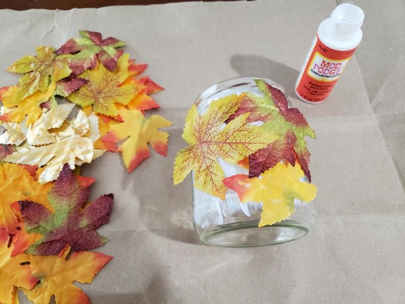 Fall leaves glued down to a glass candle holder, with extra leaves and a container of mod podge on a table