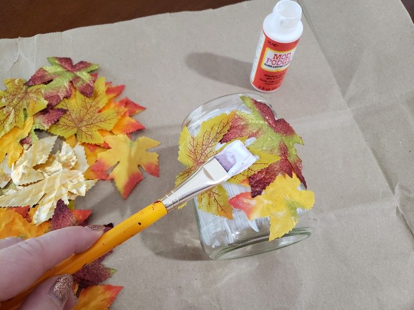 A paintbrush being used to apply mod podge to a glass candle holder covered in fall leaves
